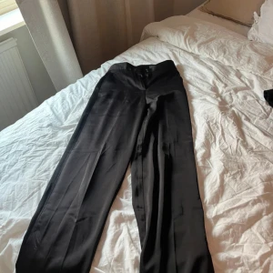 Svarta kostymbyxor - Snygga svarta kostymbyxor med hög midja och raka ben. Perfekta för en stilren look. Byxorna har en diskret bakficka och stängs med dragkedja och knapp framtill. Slits nere vid byxorna 