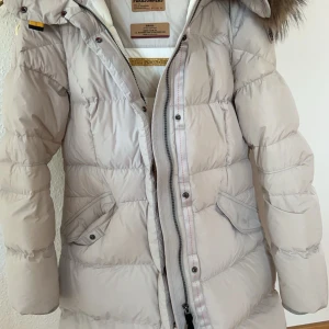 Parajumper Jacka - Snygg och varm vinterjacka med huva och pälskant. Jackan har en smal passform och är tillverkad i ett lätt och mjukt material. Den har två fickor framtill och en innerficka. Jackan är i gott skick och har endast använts ett fåtal gånger