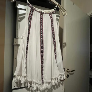 Coachella H&M Vit topp etniskt mönster - Säljer ny vit ärmlös topp från H&M Coachella. Den har ett coolt etniskt mönster i svart och rosa längs framsidan och runt halsen. Toppen är dekorerad med söta tofsar längst ner, vilket ger den en bohemisk känsla. Perfekt för sommar eller festivaler