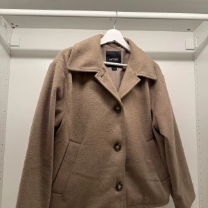Beige jacka från Monki - Säljer en superfin beige jacka från Monki i storlek S. Den har en klassisk design med stora knappar och en mjuk, mysig känsla. Perfekt för höst och vår! Jackan har långa ärmar och två praktiska fickor framtill.