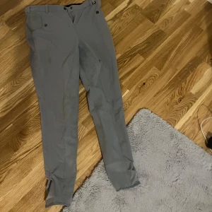 Grå ridbyxor från Jacson - Säljer ett par grå ridbyxor från Jacson i storlek 170. De har en snygg och praktisk design med knappar och dragkedja framtill. Perfekta för ridning och har en bekväm passform. Byxorna är i bra skick och redo för nya äventyr i stallet!