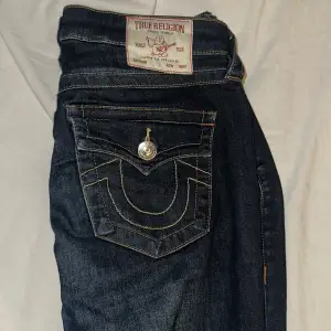 Säljer ett par snygga True Religion jeans i mörkblå denim. De har en klassisk bootcut-stil och är midwaist, vilket ger en bekväm passform. Jeansen har de ikoniska bakfickorna med knappar och kontrastsömmar som verkligen sticker ut. Perfekta för både vardag och fest!