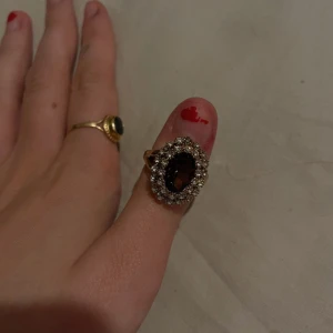 Guldfärgad ring med stor mörk sten - Säljer en elegant guldfärgad ring med en stor mörk sten i mitten, omgiven av små gnistrande stenar. Perfekt för att ge en lyxig touch till din outfit. Ringen är i nyskick och passar bra till både vardag och fest. 💍✨