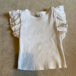 Vit ribbad topp från Zara - Säljer en söt vit ribbad topp från Zara med volangdetaljer på ärmarna. Perfekt för en gullig stil. Toppen är i storlek S och passar utmärkt till både jeans och kjol.