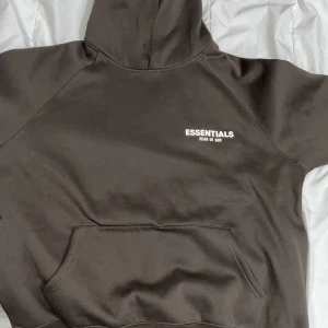 Brun hoodie från Essentials Fear of God - Säljer en snygg brun hoodie från Essentials Fear of God. Den har en klassisk design med en känguruficka och textlogga på bröstet och ärmen. Perfekt för en stilren och avslappnad look.