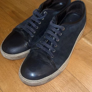 Marinblå/svart sneakers från Lanvin - Snygga svarta sneakers från Lanvin med snörning och en stilren design. Skorna har en gummisula och är tillverkade i skinn och mocka, vilket ger en lyxig känsla. Storlek 45 men passar 44, kvitto finns