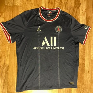 Nike Jordan PSG xl t-shirt säsong 21/22 - Snygg mörkblå PSG fotbollströja med korta ärmar och röd-vita detaljer vid krage och ärmslut. Tröjan har Jordan-logga och PSG-emblem på bröstet samt texten 'Accor Live Limitless' på framsidan. Perfekt för fotbollsfans!