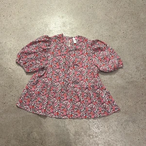 Blommig blus från Vero Moda strl 146 - En  söt blommig blus från Vero Moda i storlek 146. Blusen har korta  puffiga ärmar. Tre rosetter i mitten av blusen. Den är i en härlig mix av rosa, orange och vit med ett blommigt mönster. Perfekt för vår och sommar!