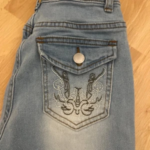 Blå bootcut jeans med broderad ficka - Snygga ljusblå bootcut jeans med en unik broderad design på bakfickan. Jeansen har en klassisk femficksmodell och en knappstängning. Perfekta för en avslappnad stil med en touch av detaljer.