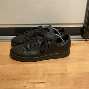 Svarta stan Smith sneakers från Adidas - Snygga helsvarta stan Smith Adidas sneakers med klassisk design. Skorna har snörning och är tillverkade i skinn. Perfekta för en stilren look.