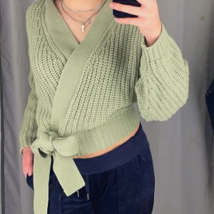 Grön stickad omlottkofta - Säljer en mysig grön stickad omlottkofta med knytband i midjan. Koftan har långa ärmar och en ribbad struktur som ger en stilren look. Perfekt för kyligare dagar.