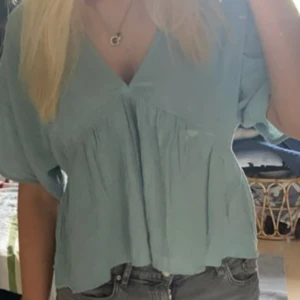 Ljusblå/turkos kort blus med v-ringning - Säljer en luftig ljusblå/turkos blus med v-ringning och korta ärmar. Blusen har en avslappnad passform och volangdetaljer som ger en romantisk känsla. Perfekt för varmare dagar. Blusen är också i linne liknande material 