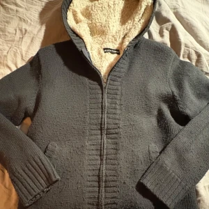 Grå stickad cardigan - Mysig grå stickad jacka från Much More med teddyfoder i huvan. Jackan har dragkedja framtill och ribbade detaljer vid ärmslut och nederkant. Perfekt för kyliga dagar.