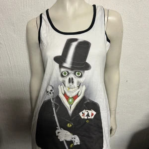 Linne från Insanity storlek S/M - Coolt vitt linne med ett unikt skelettmotiv. Skelettet bär en hög hatt och håller en käpp med dödskalle. Perfekt för dig som vill sticka ut med en edgy stil.