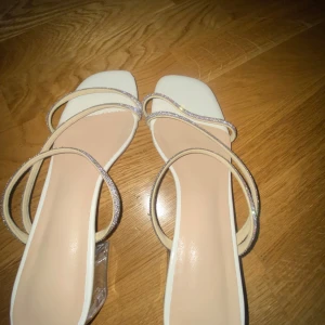 Vita sandaler med glittriga remmar - Snygga vita sandaler med glittriga remmar och transparent klack. Perfekta för att ge en touch av glamour till din outfit. De har en elegant design med flera remmar som korsar foten. Pris kan diskuteras!