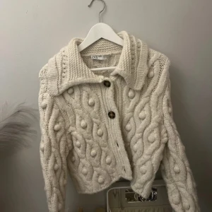 Vit stickad kofta från Zara - Säljer en mysig vit stickad kofta från Zara med ett unikt flätmönster och stora knappar. Koftan har en bred krage och långa ärmar, perfekt för kyliga dagar. Den är både stilren och bekväm.