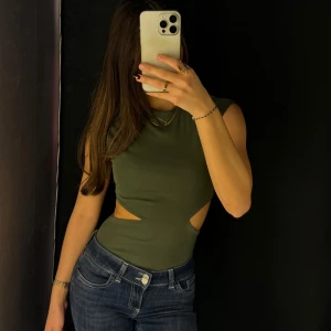 Mörkgrön body - Snygg mörkgrön body med cut-out detaljer i sidorna. Toppen är ärmlös och har en tight passform som framhäver figuren. Perfekt för en trendig look. Den är i storlek XS men passar även en S.
