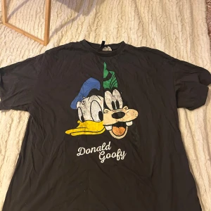 Svart t-shirt med Donald och Goofy från Disney - Cool svart t-shirt med tryck av Donald och Goofy i färg på framsidan. Perfekt för alla Disney-fans som vill ha en avslappnad stil. T-shirten har en rund hals och korta ärmar.
