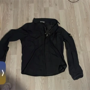 Svart jacka från Stone Island - Säljer en stilren svart jacka från Stone Island. Jackan har långa ärmar och en klassisk krage. Den är försedd med knappar och har det ikoniska Stone Island-märket på ärmen. Perfekt för dig som vill ha en tidlös och snygg look.