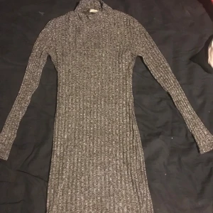Grå ribbad klänning från Kikiriki - Säljer en stilren grå ribbad klänning från Kikiriki i storlek XS. Klänningen har en tight passform och långa ärmar, perfekt för kyligare dagar. Den höga kragen ger en elegant touch. Passar utmärkt för både vardag och speciella tillfällen.