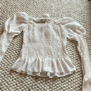 Vit blus från Boohoo - Säljer en söt vit blus från Boohoo med smockdetaljer och volangkant. Blusen har långa ärmar och en knytning i ryggen. Perfekt för en romantisk look!