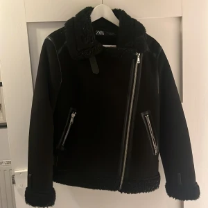 Svart jacka från Zara - Snygg svart jacka från Zara med dragkedja och pälsdetaljer. Jackan har långa ärmar och en cool krage. Perfekt för att hålla sig varm och stilren under kyliga dagar.
