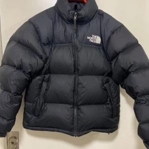 Svart dunjacka från The North Face - Säljer en svart pufferjacka från The North Face. Den är perfekt för kalla dagar och har en klassisk design med logotyp på bröstet och ryggen. Inbyggd luva finns i kragen. Modellen heter 1996 Retro Nupste Jacket. Passar både kille och tjej. Denna jackan är i absolut nyskick, endast använd 2-3 ggr. Sonen fick en ny i julklapp och kommer därför inte använda denna mer. Nypris: 3895:- på Zalando. Mitt pris kan diskuteras vid snabb affär! 