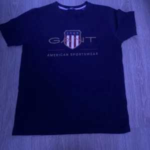 Svart t-shirt från Gant - Säljer en svart t-shirt från Gant med tryck på framsidan. T-shirten har en klassisk rund hals och korta ärmar. Perfekt för en avslappnad stil.