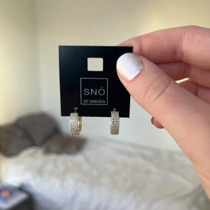 Guldglittriga örhängen från SNÖ of Sweden - Snygga guldglittriga örhängen från SNÖ of Sweden. Dessa eleganta örhängen har en glittrig yta som fångar ljuset perfekt. Perfekta för att ge en touch av glamour till din outfit.