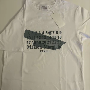 Vit t-shirt från Maison Margiela - Snygg vit t-shirt från Maison Margiela med deras ikoniska siffertryck i svart på framsidan. Perfekt för en stilren och modern look. T-shirten har en normal passform och är kortärmad.