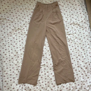 Beige kostymbyxor från Bik Bok - Snygga beige kostymbyxor från Bik Bok i storlek 34. Byxorna har en klassisk design med hög midja och raka ben. De är utrustade med dragkedja och knapp framtill för en säker passform.