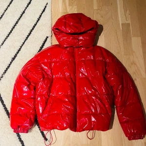 Röd glansig pufferjacka - Säljer en cool röd pufferjacka med glansig finish. Jackan har en dragkedja framtill och en mysig huva. Perfekt för att hålla värmen under kyliga dagar. Den har en modern look som verkligen sticker ut!