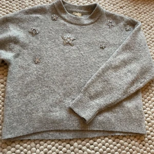 Grå stickad tröja med stjärndetaljer från H&M - Säljer en mysig grå tröja från H&M med glittriga stjärndetaljer på framsidan. Tröjan har långa ärmar och ribbade muddar vid ärmslut och nederkant. Perfekt för kyliga dagar!