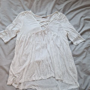Vit blus  - Säljer en luftig vit blus med v-ringad design och snygg snörning framtill. Blusen har trekvartsärmar och ett avslappnat fall. Perfekt att ha över t.ex. över bikinin
