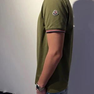 Olivgrön t-shirt från Moncler - Snygg olivgrön t-shirt från Moncler med korta ärmar och en liten logga på ärmen. T-shirten har ribbade kanter med röd och vit detaljering. Moddellen är 173 och väger runt 60kg! 💯Skick 8/10 bara lite fläckar i armhålan men inget som är så synligt👌❗️OBS osäker om autenticitet ❗️vid fler frågor kom privat! 💯👍