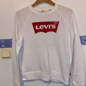 Vit sweatshirt från Levi's - Säljer en vit sweatshirt från Levi's med röd logga på bröstet. Tröjan har långa ärmar och en klassisk rund halsringning. Perfekt för en avslappnad stil.