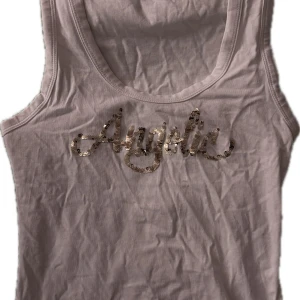Vit linne topp med paljetter - Säljer en vit ärmlös topp med texten 'Angelic' i glittrande paljetter på framsidan. Perfekt för en glittrig look!