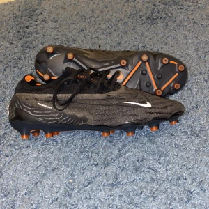 Svarta fotbollsskor från Nike - Snygga svarta fotbollsskor från Nike med orange detaljer och snörning. Skorna har en modern design med mönstrad yta och är perfekta för fotbollsplanen. Skorna var köpta 2022 på Inter sport och var förstora men har nu slutat på fotboll. Det är i pirincip helt oanvända. 