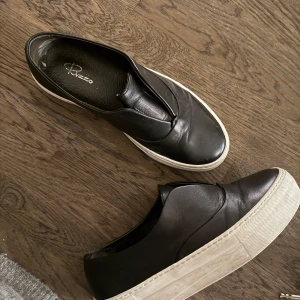 Svarta slip-on sneakers från Rizzo - Snygga svarta slip-on sneakers från Rizzo i skinn med vit sula. Tveka inte att höra av dig!🥰