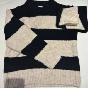 Randig stickad tröja - Mysig stickad tröja med breda ränder i svart och beige. Tröjan har långa ärmar och en klassisk passform, perfekt för kyligare dagar.
