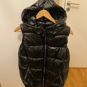 Svart pufferväst med huva - Snygg svart pufferjacka med huva och dragkedja framtill. Jackan har en glansig finish och är perfekt för kyligare dagar. Den är ärmlös och har en modern look.