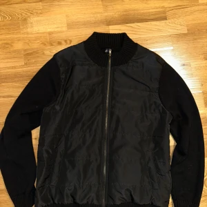 Cardigan jacka - Snygg svart cardigan jacka med dragkedja och ribbad krage. Jackan har långa ärmar och en stilren design som passar perfekt för höst och vår. Materialet verkar vara en blandning av syntet och stickat, vilket ger en unik look. Helt ny, storlek L men passar M med