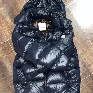  Moncler maya  - Säljer en snygg svart dunjacka från Moncler med glansig finish. Jackan har en huva och dragkedja framtill. Den är quiltad för extra värme och har Monclers logga på ärmen. Perfekt för kalla dagar! Liten skada på armen men kan täckas över. Pm för bild 