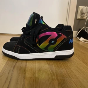 Svarta Heelys med regnbågsdetaljer - Säljer ett par svarta Heelys med regnbågsfärgade detaljer och snörning. Skorna har en hjulmekanism i sulan för rullning. Perfekta för den som vill glida fram med stil! Använt två gånger❤️