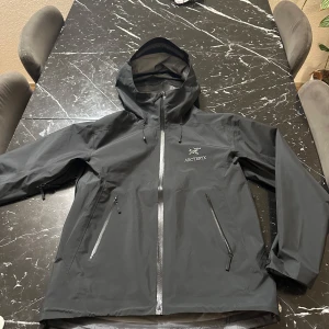 Svart skaljacka från Arc'teryx - Säljer en svart skaljacka från Arc'teryx i Gore-Tex-material. Jackan har en justerbar huva och flera dragkedjeförsedda fickor. Perfekt för utomhusaktiviteter med sin vattentäta design.