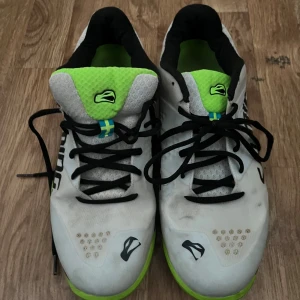 Vitas och gröna träningsskor från Salming - Säljer ett par vita träningsskor från Salming med svarta och gröna detaljer. Skorna har snörning och en sportig design med Salming-logga på sidan. Perfekta för inomhusträning eller löpning.