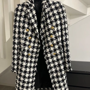 Svartvit rutig kappa med guldknappar - Snygg svartvit rutig kappa med dubbelknäppning och stiliga guldknappar. Perfekt för att ge en elegant touch till din outfit. Klassisk design som aldrig går ur tiden.