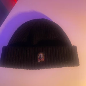 Svart ribbad mössa från Parajumpers - Snygg svart ribbad mössa från Parajumpers med ett broderat märke framtill. Perfekt för att hålla värmen under kyliga dagar. Mössan har en klassisk design och är tillverkad i ett mjukt material.