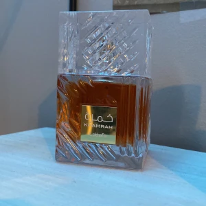 Kamrah Parfym från Lattafa - Elegant parfymflaska från Lattafa med namnet Kamrah. Flaskan har en unik design med ett mönstrat, genomskinligt lock och en fyrkantig form. Vätskan inuti är bärnstensfärgad, vilket ger en lyxig känsla. Perfekt för den som söker en distinkt och stilfull doft.