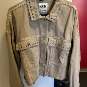 Beige jeansjacka från Zara - Säljer en beige jeansjacka från Zara med coola nitar på kragen och fickorna. Jackan är croppad och har en avslappnad passform med knappar framtill. Perfekt för en trendig look!